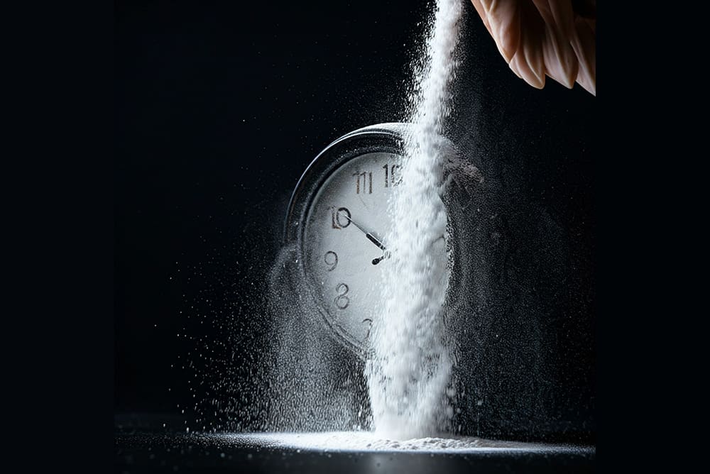 A hand pours a stream of white powder over a clock, with scattered powder on the surface.